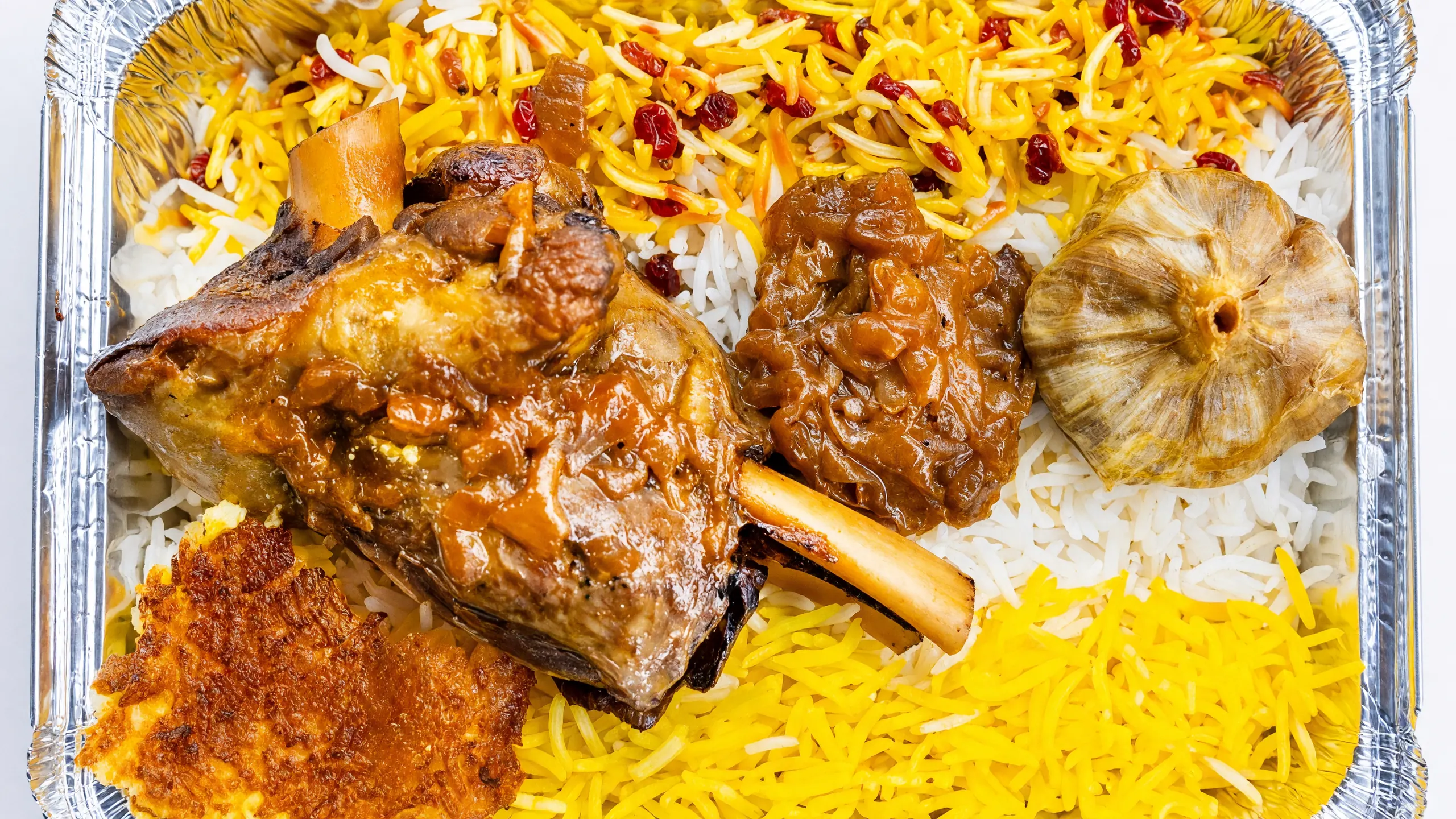 Braised lamb shank in tomato sauce with a roasted garlic bulb, saffron rice with barberries and crispy tahdig in a foil tray at AFRA Grill, Saskatoon