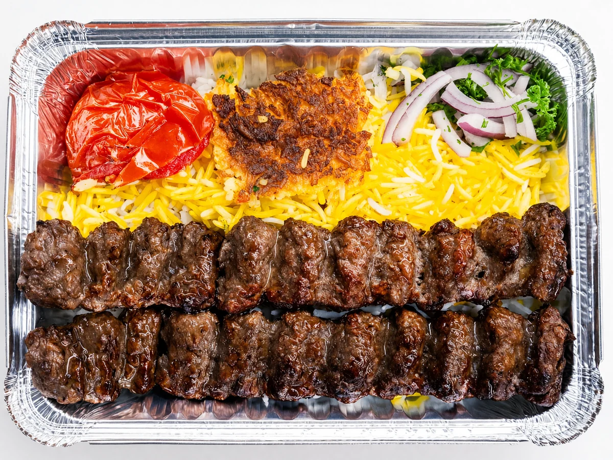 Two skewers of grilled ground beef koobideh kabab on saffron rice with tahdig, a grilled tomato and sliced red onion in a foil tray at AFRA Grill, Saskatoon